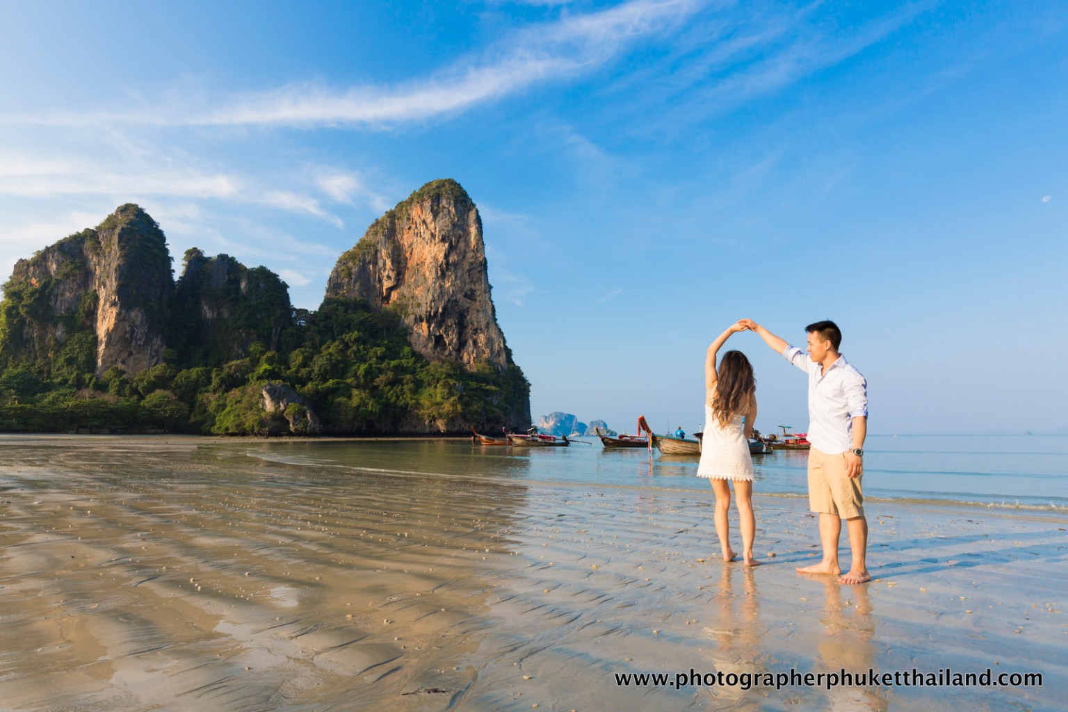 A couple enjoying a romantic moment on the beach in Krabi, Thailand, with limestone cliffs in the background and traditional boats in the water.