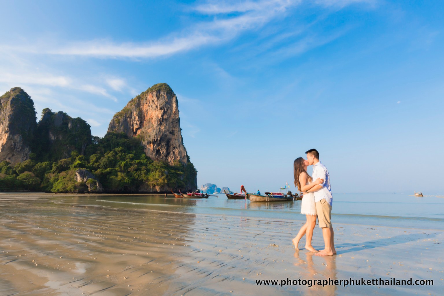 A couple sharing a kiss on a beach with rocky cliffs and boats in the background under a clear blue sky.