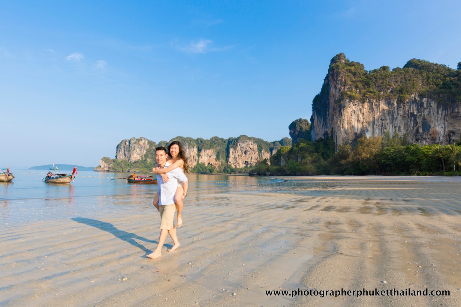A couple enjoying a playful moment by the beach, with the man giving the woman a piggyback ride. In the background, there's a serene beach landscape with clear blue sky and rocky cliffs.