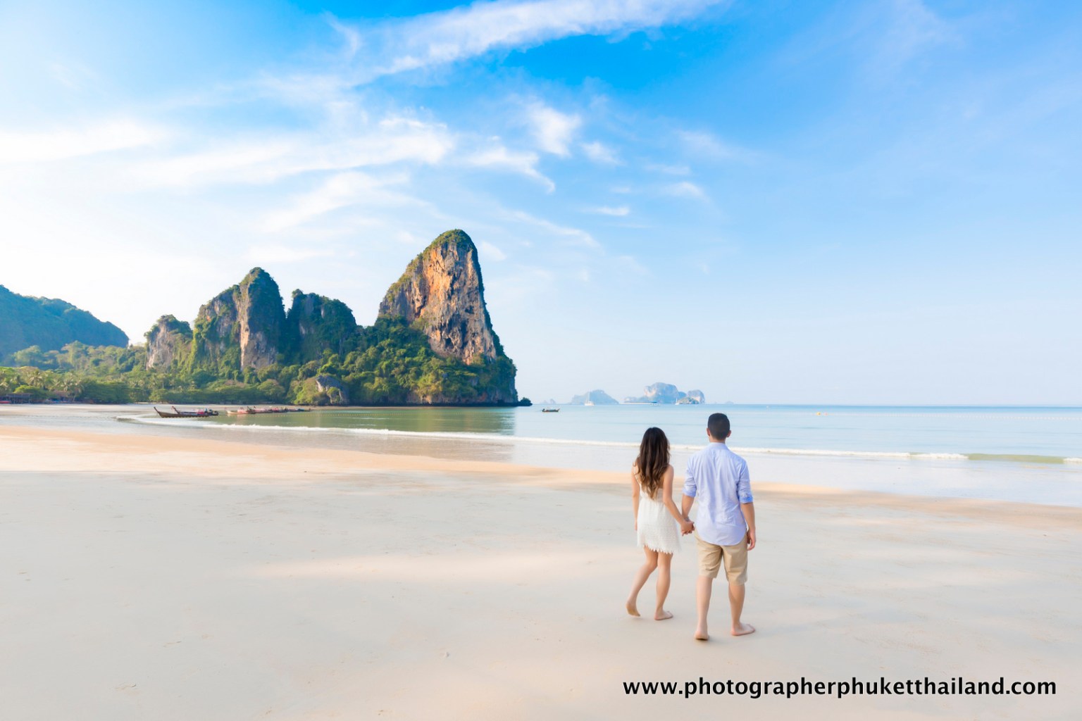 A couple walking hand in hand on a sandy beach with rocky cliffs and a clear blue sky in the background.