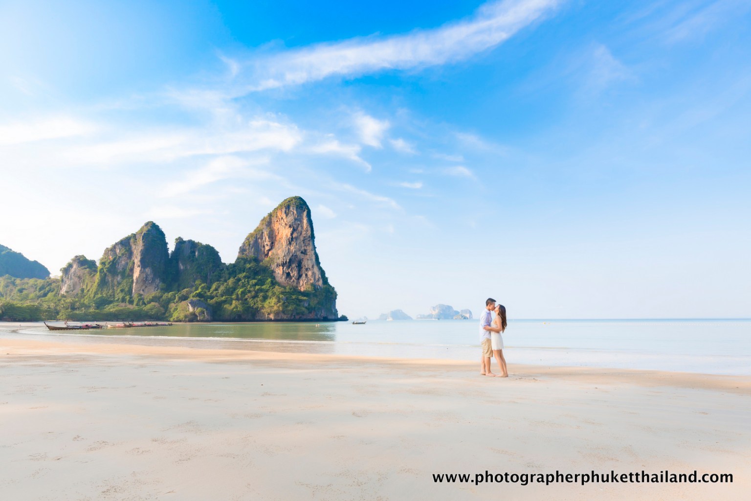 A couple embracing on a serene beach with mountains in the background and a clear blue sky.