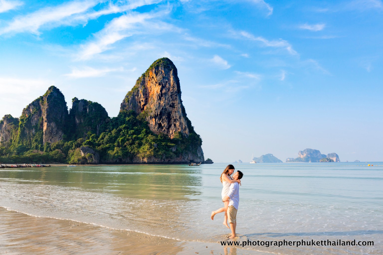 A couple enjoys a romantic moment on the beach at Krabi, Thailand, with dramatic limestone cliffs and a clear blue sky in the background.