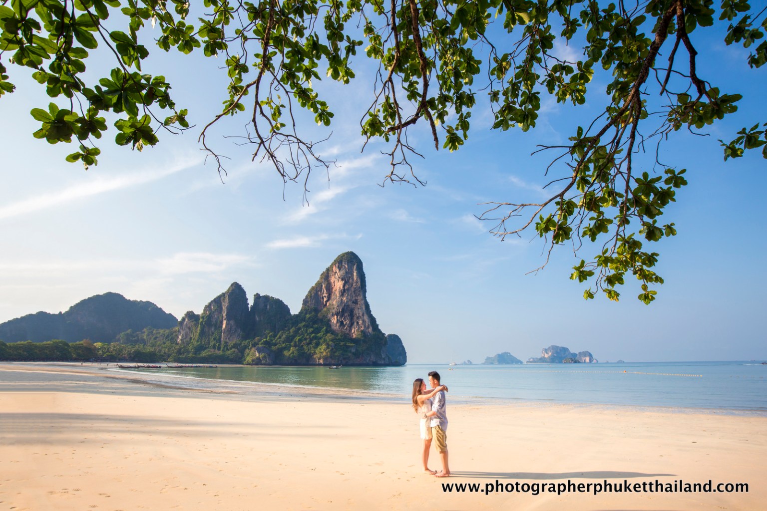 A couple embracing on a sandy beach with lush green foliage in the foreground and scenic mountains in the background under a clear blue sky.