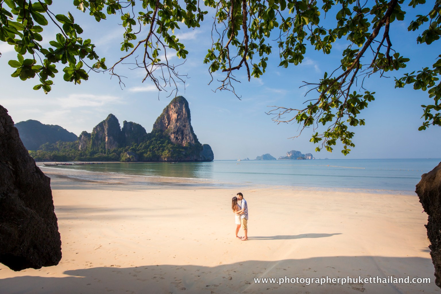 A couple embracing on a sandy beach with limestone cliffs in the background, surrounded by greenery and clear blue waters.