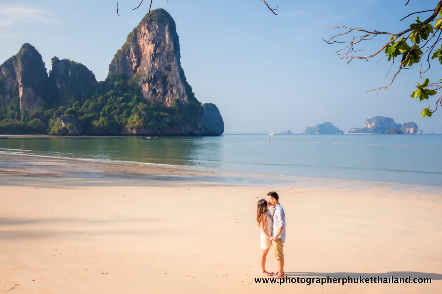 A couple holding hands on a sandy beach in Krabi, Thailand, with limestone cliffs and islands in the background.