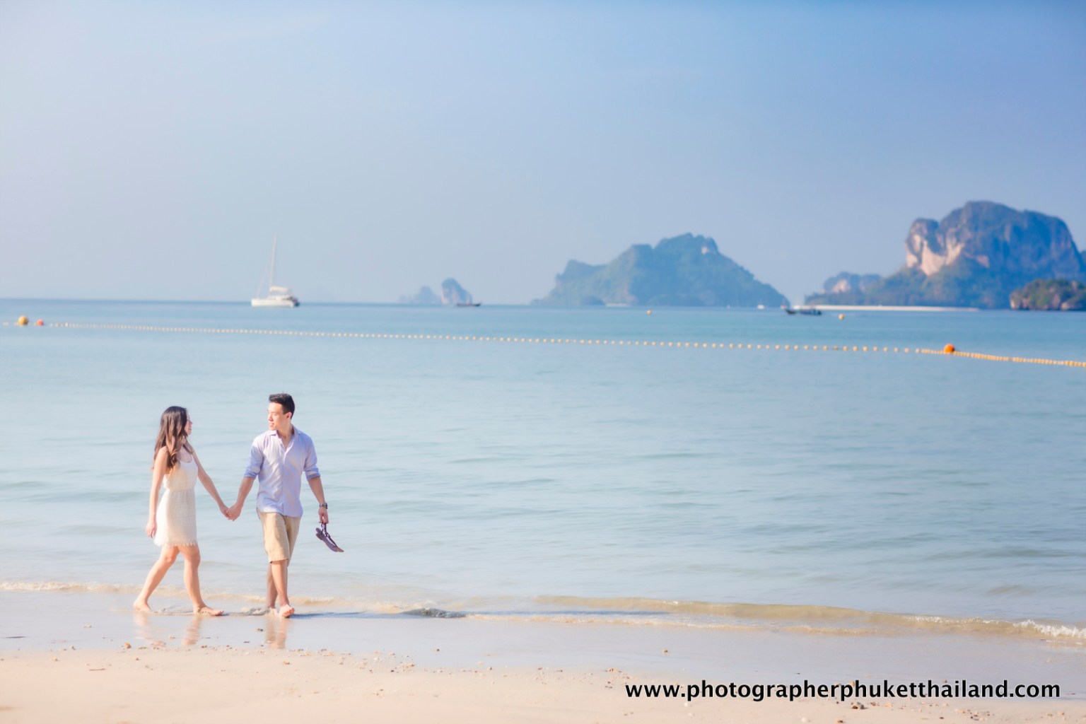 Honeymoon couple walking hand in hand on the beach in Krabi, Thailand, with limestone cliffs in the background.