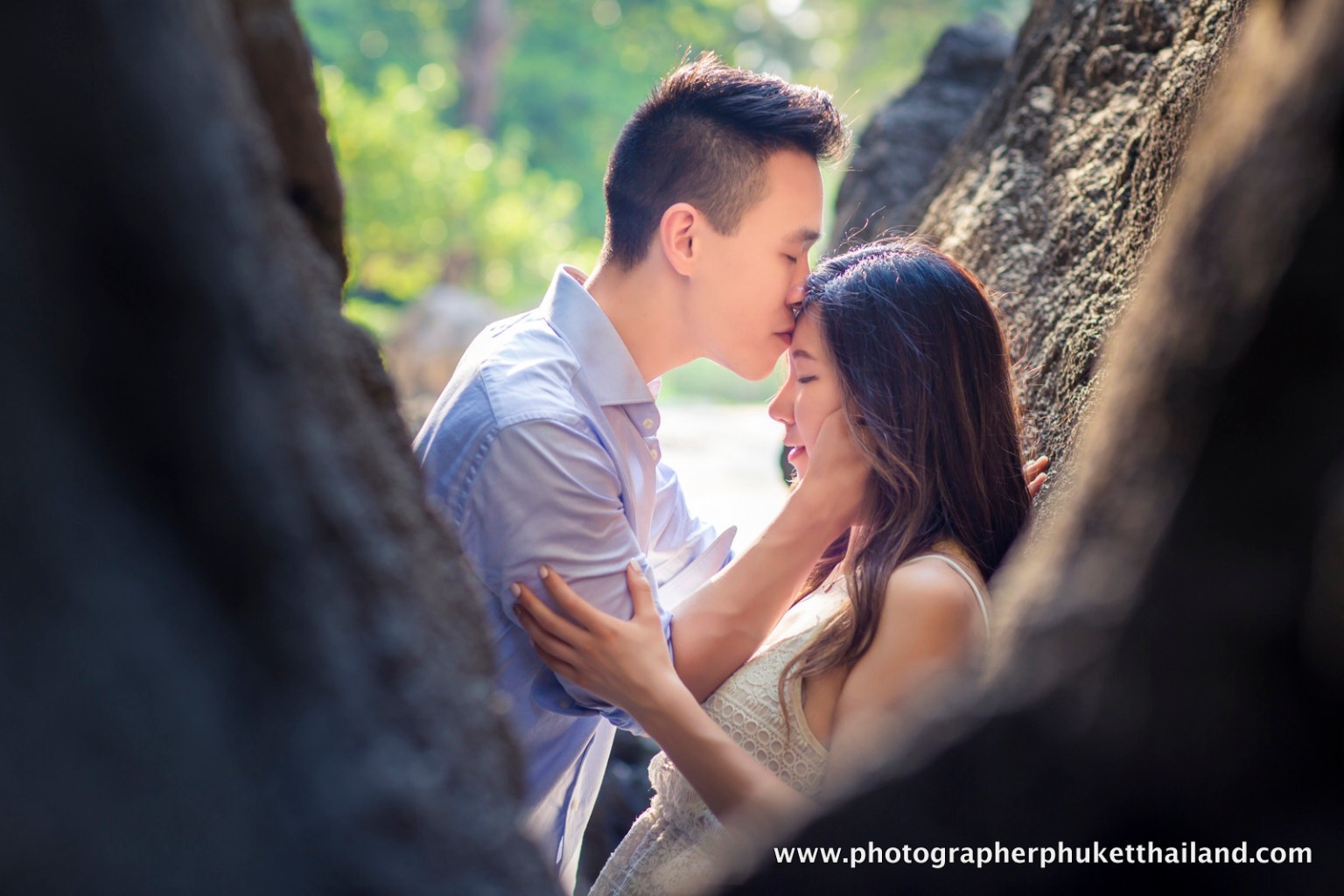 A couple sharing an intimate moment, with the man gently kissing the woman's forehead while she holds his face, framed by natural rock formations and soft green foliage in the background.