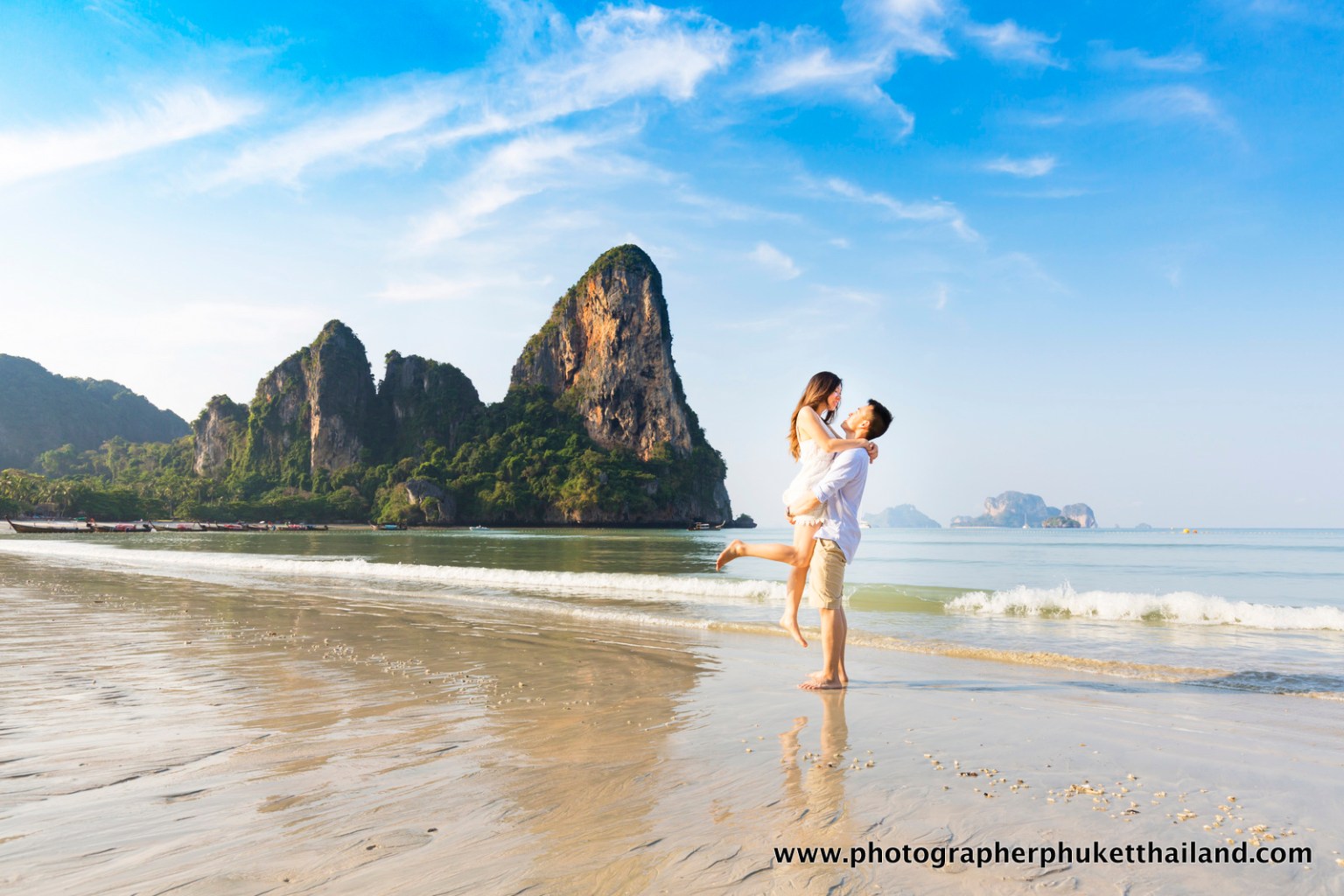 A couple embracing on a beach with limestone cliffs in the background and a clear blue sky.