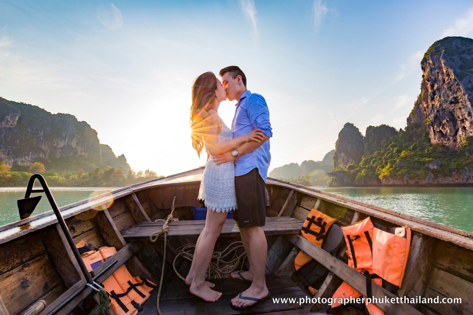 A couple sharing a romantic kiss on a boat with a scenic backdrop of limestone cliffs and water in Krabi, Thailand, during sunset.