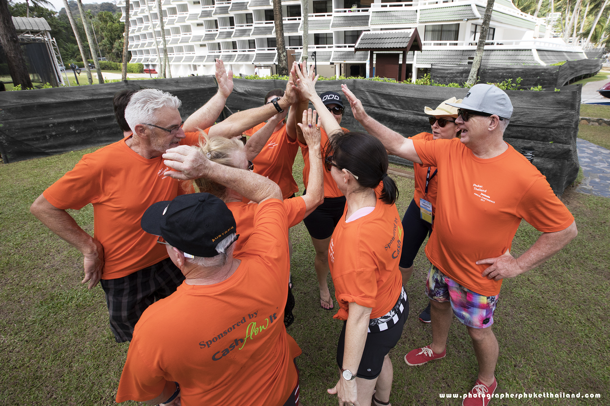 corperate A group of people wearing orange t-shirts is gathered outdoors, celebrating and giving high-fives with enthusiasm. The background features greenery and a building.