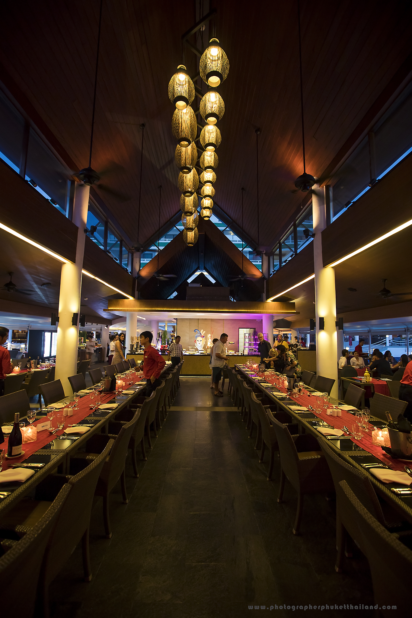 Interior of a restaurant set for an event, featuring long tables with red tablecloths, elegant lighting fixtures, and people preparing for the dinner.