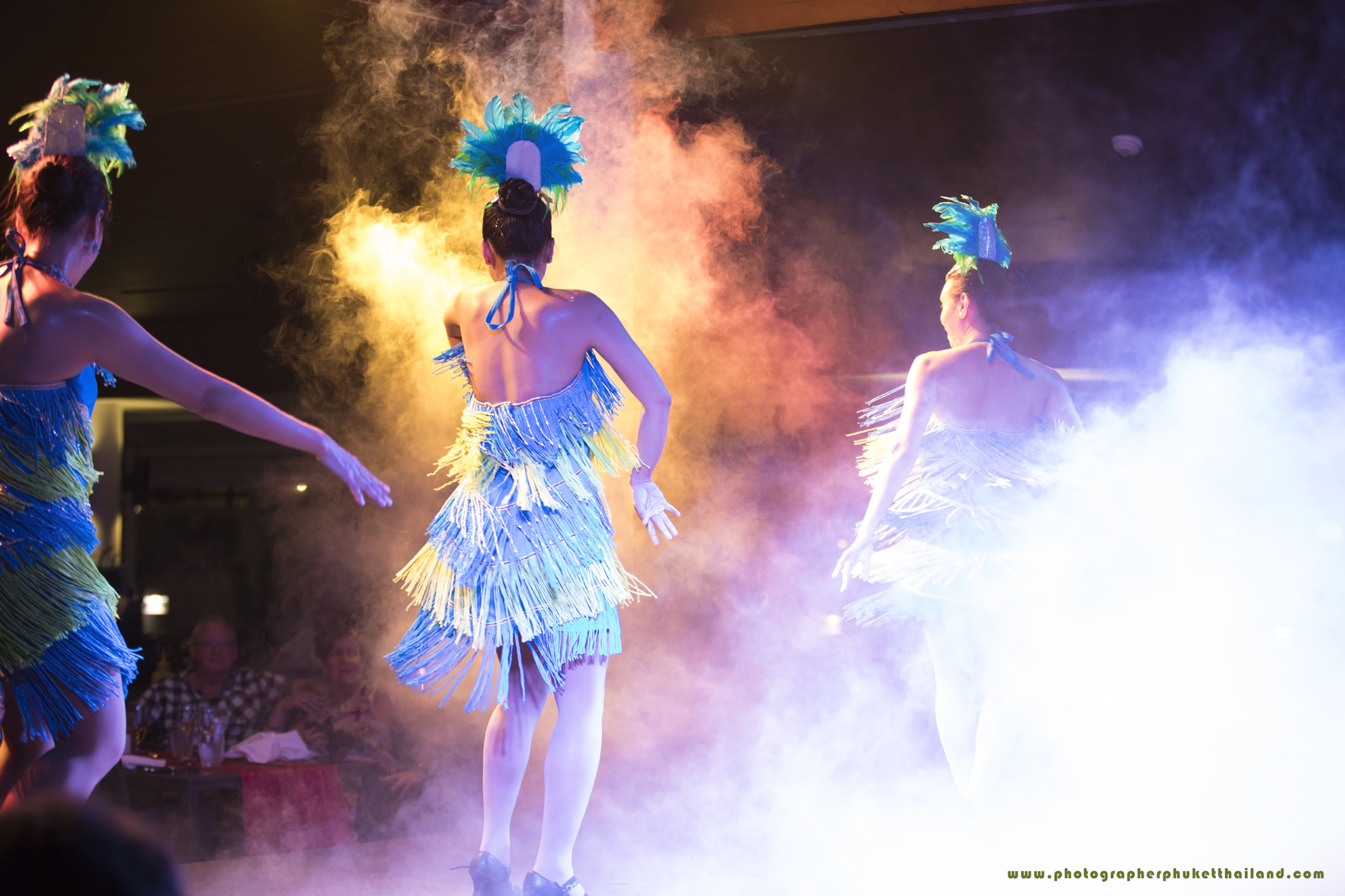 Three dancers in colorful fringe costumes performing on stage in after wedding party surrounded by colorful smoke effects.