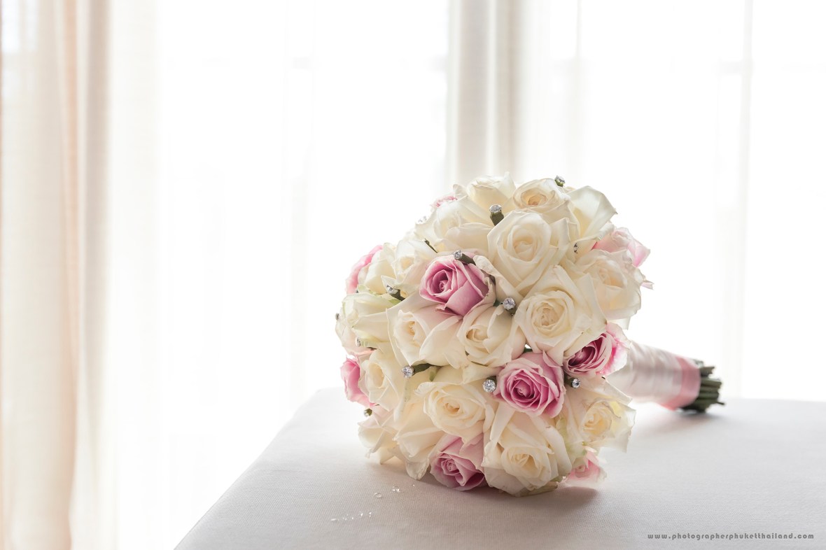 A close-up of a wedding bouquet featuring white and pink roses, adorned with decorative elements, positioned on a table near a window.
