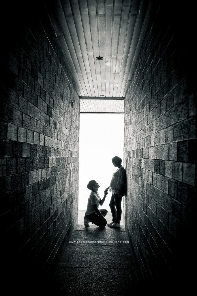 A black and white photograph of a couple in a narrow corridor, with one person kneeling and offering a gesture to the other, who stands facing them.