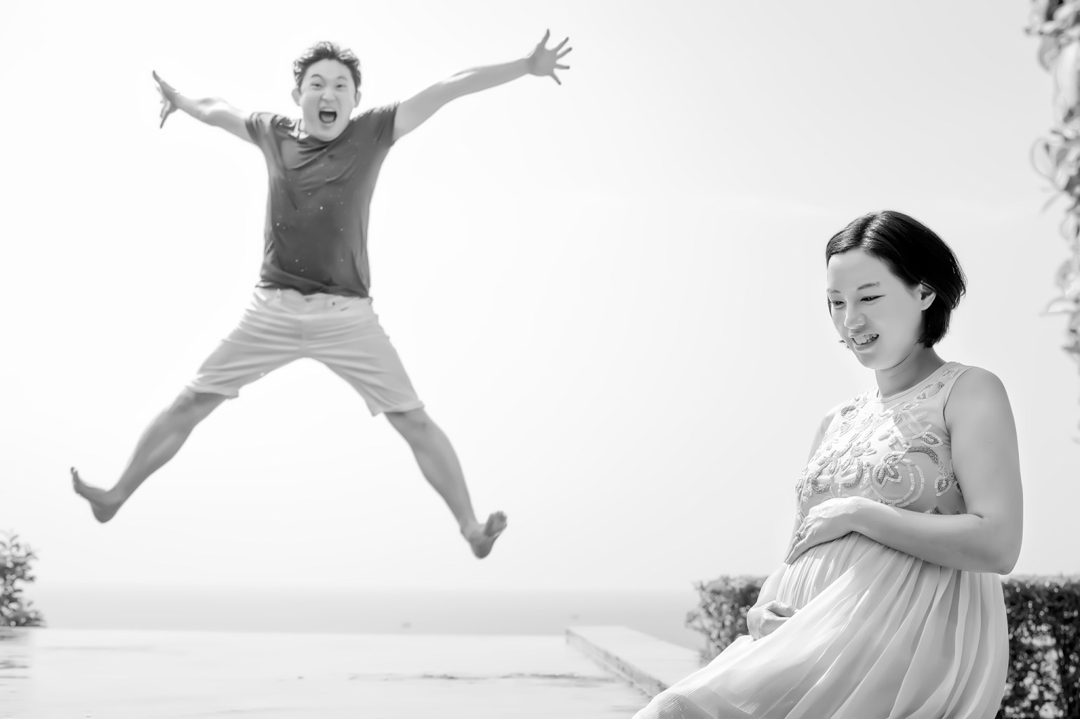 A playful man jumping joyfully in the air beside a pregnant woman, who is smiling and gently cradling her baby bump. The photo is in black and white, capturing a moment of happiness.