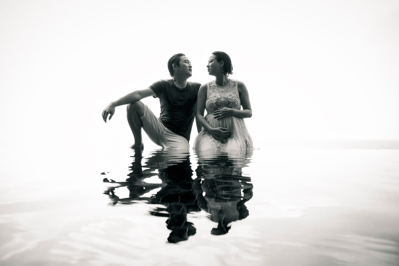 A couple sitting in shallow water, engaging in a loving conversation, captured in black and white. The man is in casual clothing while the woman is wearing a flowing, light-colored dress.