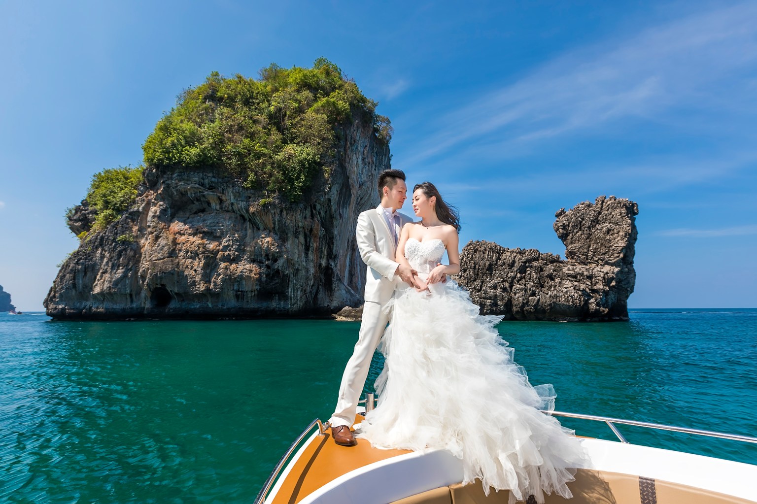 A couple in formal wedding attire stands on the bow of a boat, with lush green cliffs and clear turquoise water in the background.