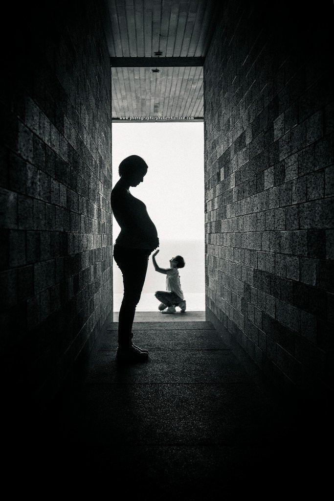 Silhouette of a pregnant woman standing in a corridor, with a child kneeling and reaching towards her in the background, creating a contrasting scene with natural light.
