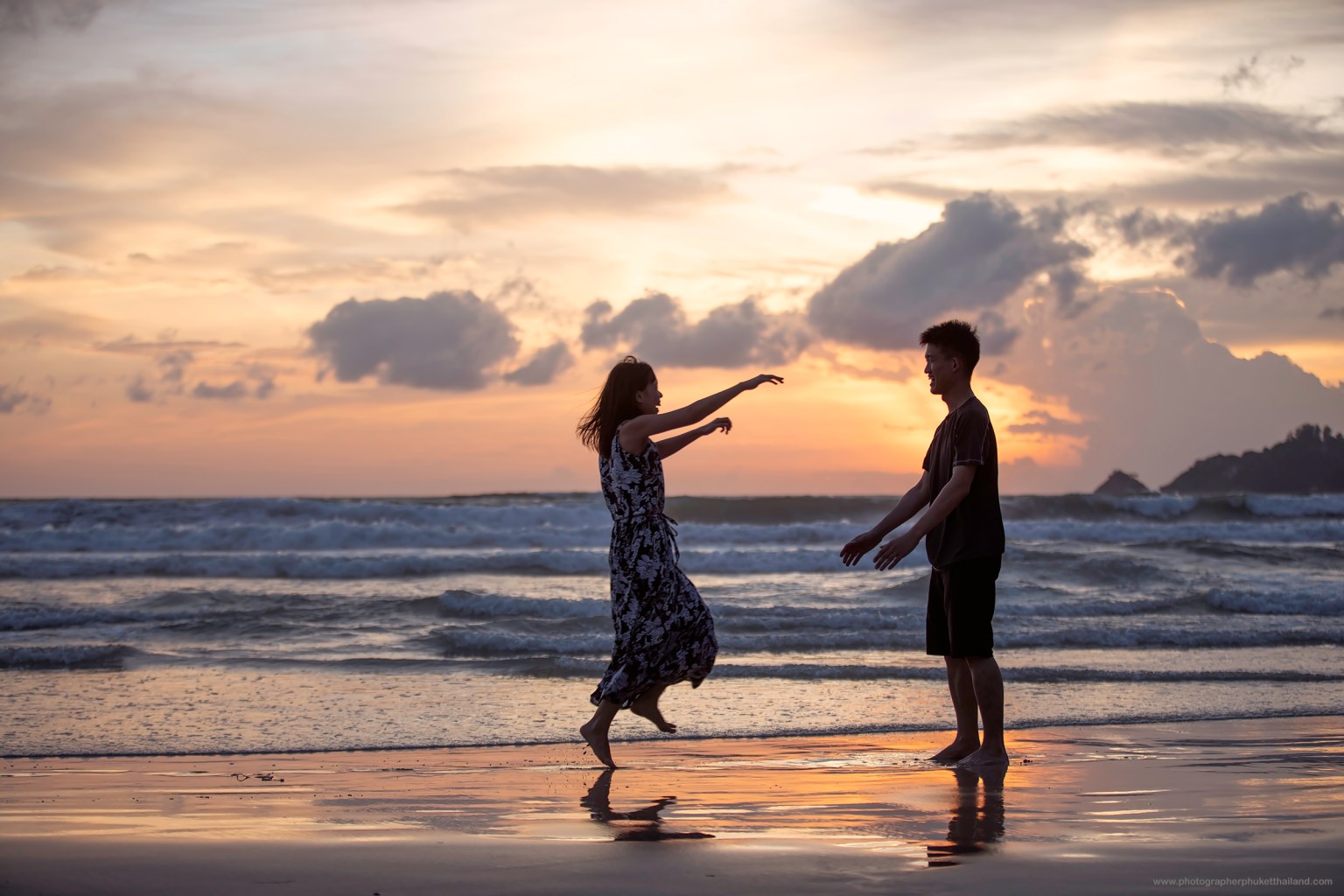 A couple joyfully playing together on the beach during sunset, with waves in the background and a colorful sky.