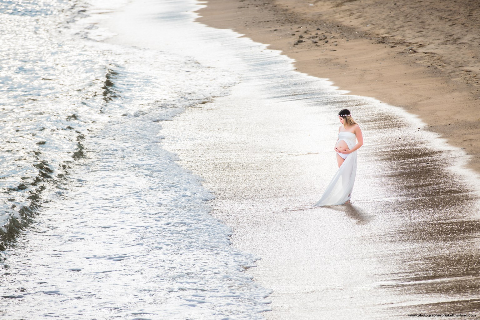 A pregnant woman in a flowing white dress stands barefoot on a sandy beach, gazing at the water as gentle waves lap at her feet. Sunlight reflects off the ocean, creating a serene and peaceful atmosphere.