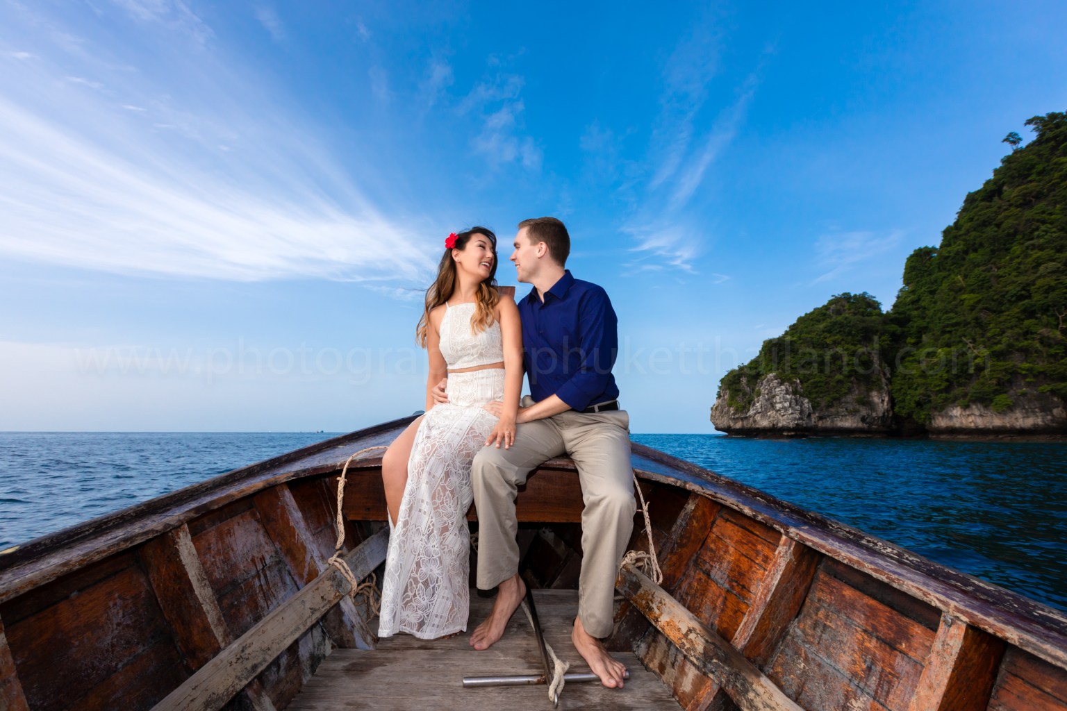 A couple sitting closely on a wooden boat in the ocean, enjoying a sunny day with a clear blue sky and distant green hills in the background.