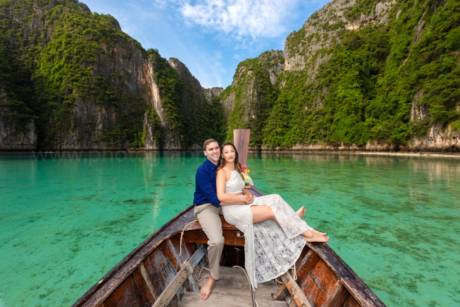 A couple sitting on a wooden boat in clear turquoise water, surrounded by lush green cliffs under a blue sky.