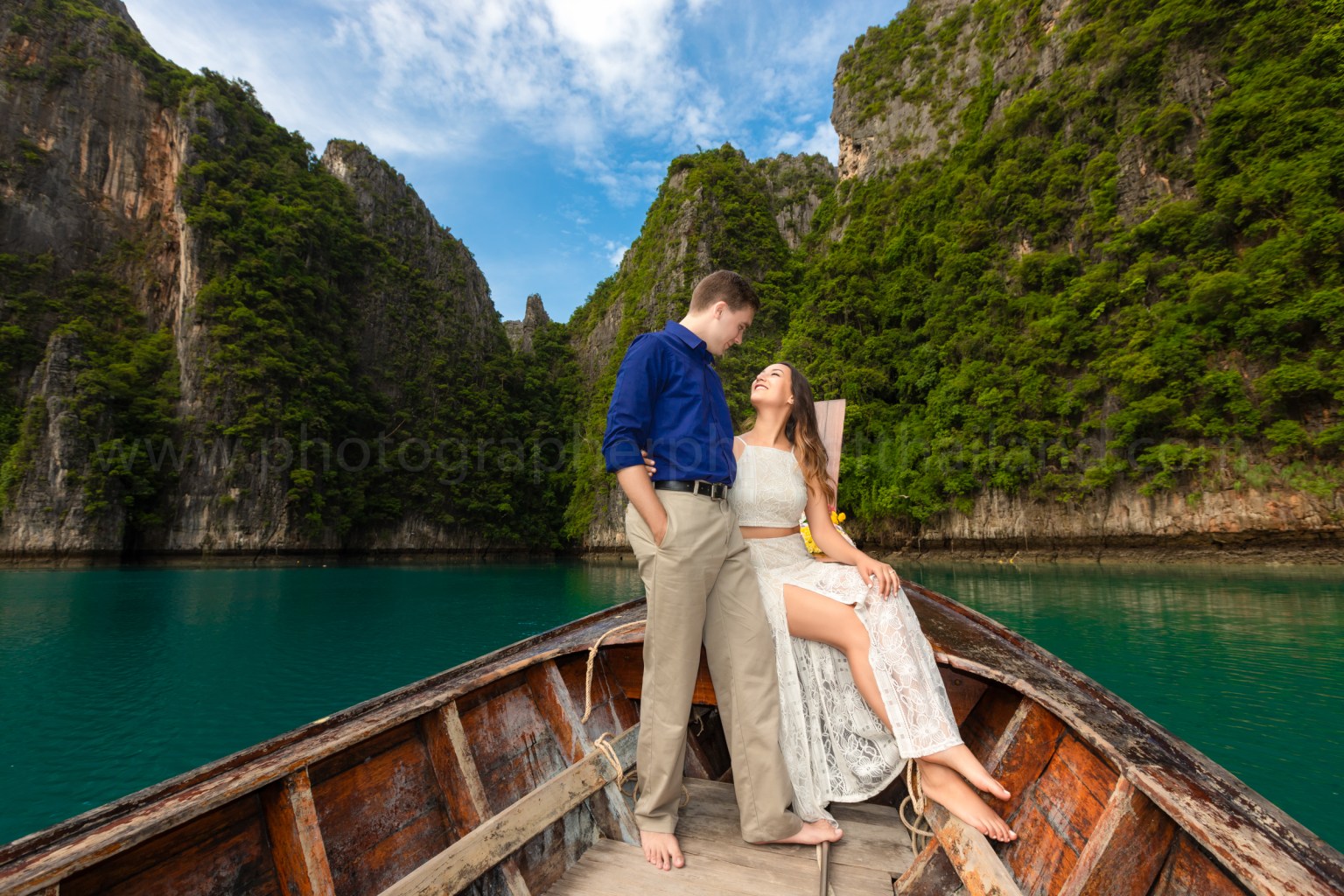 A couple joyfully posing on a boat surrounded by lush green mountains and clear turquoise waters.