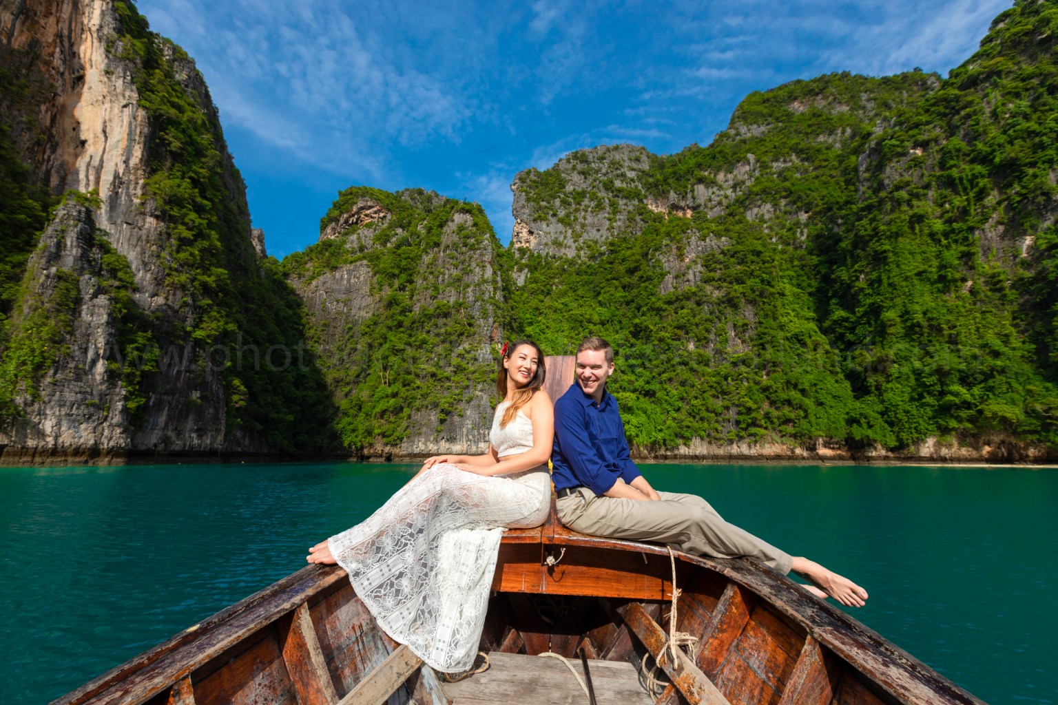 A couple sitting on the edge of a wooden boat, surrounded by lush green cliffs and blue water under a clear sky.