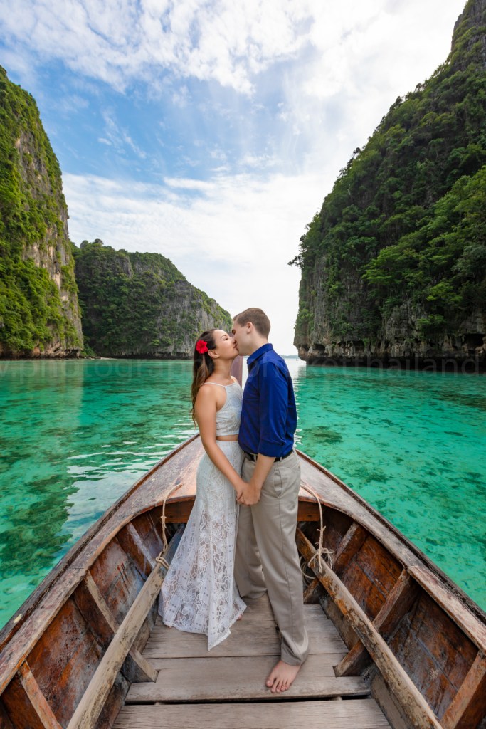 A couple standing on a wooden boat, kissing with beautiful green cliffs in the background and clear turquoise water.