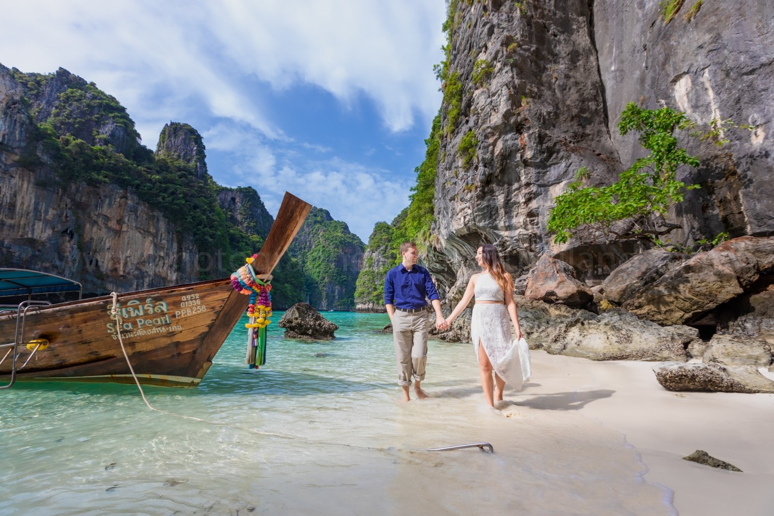 A couple walking hand in hand along a sandy beach, with a traditional wooden boat anchored nearby and lush green cliffs in the background under a partly cloudy sky.