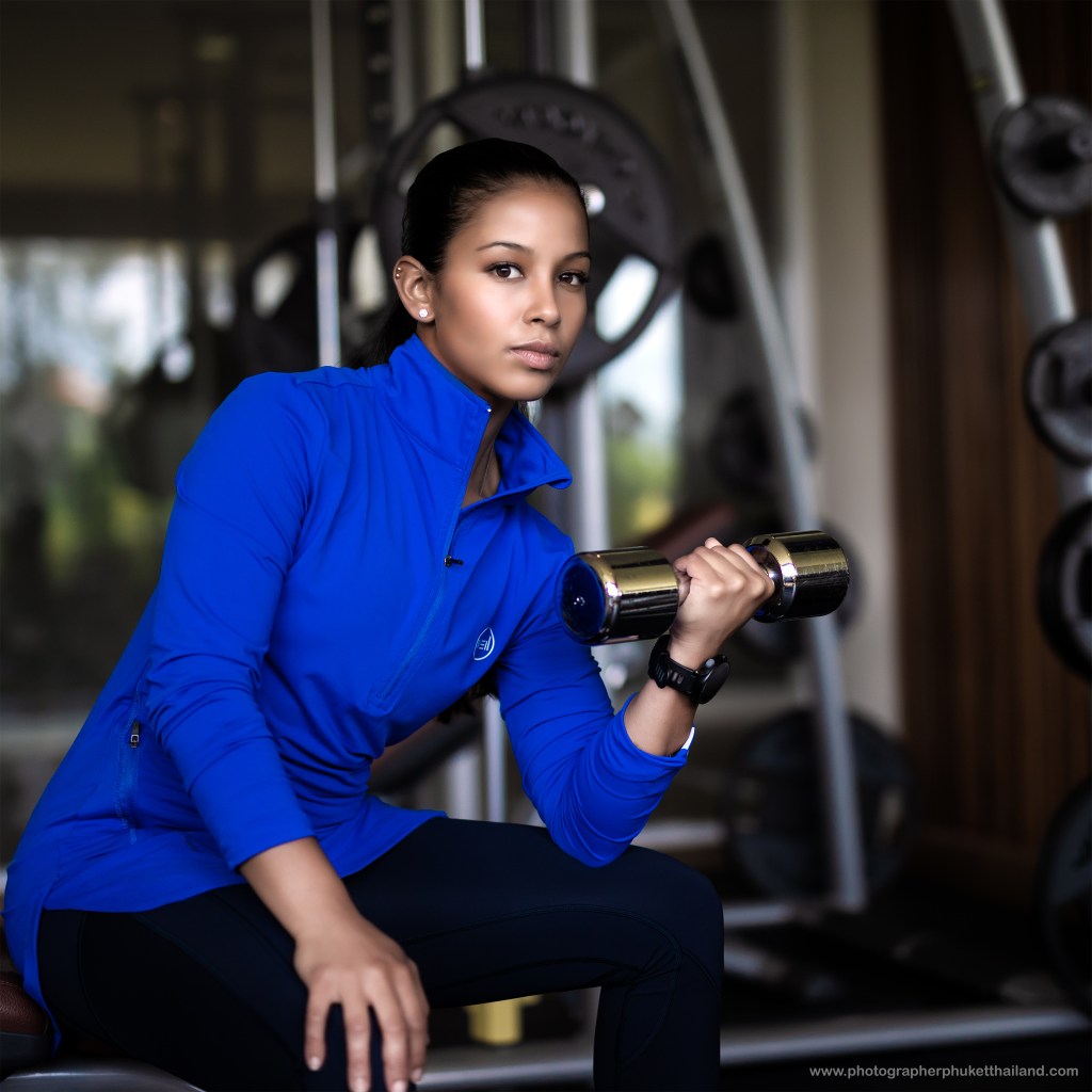 A woman in a blue athletic jacket holds a dumbbell while sitting in a gym, showcasing a focused expression.