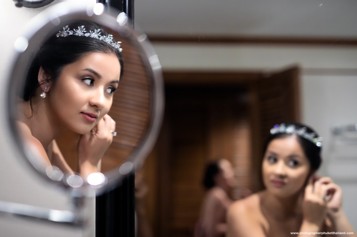 A bride adjusts her earrings in front of a mirror, showcasing her elegant hairstyle adorned with a tiara.