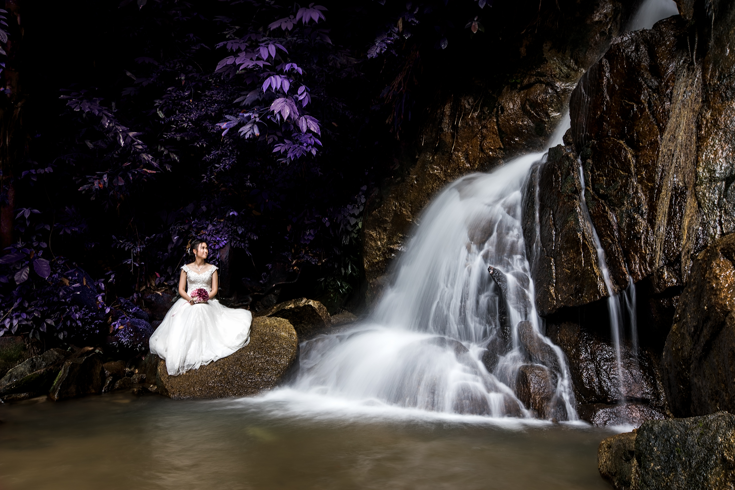 A bride in a white gown sitting on a rock by a flowing waterfall, surrounded by lush purple foliage.