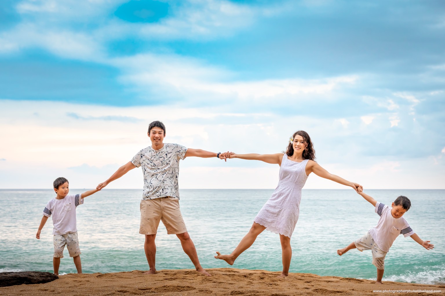 A family of four playing at the beach, holding hands and smiling, with a calm ocean and cloudy sky in the background.