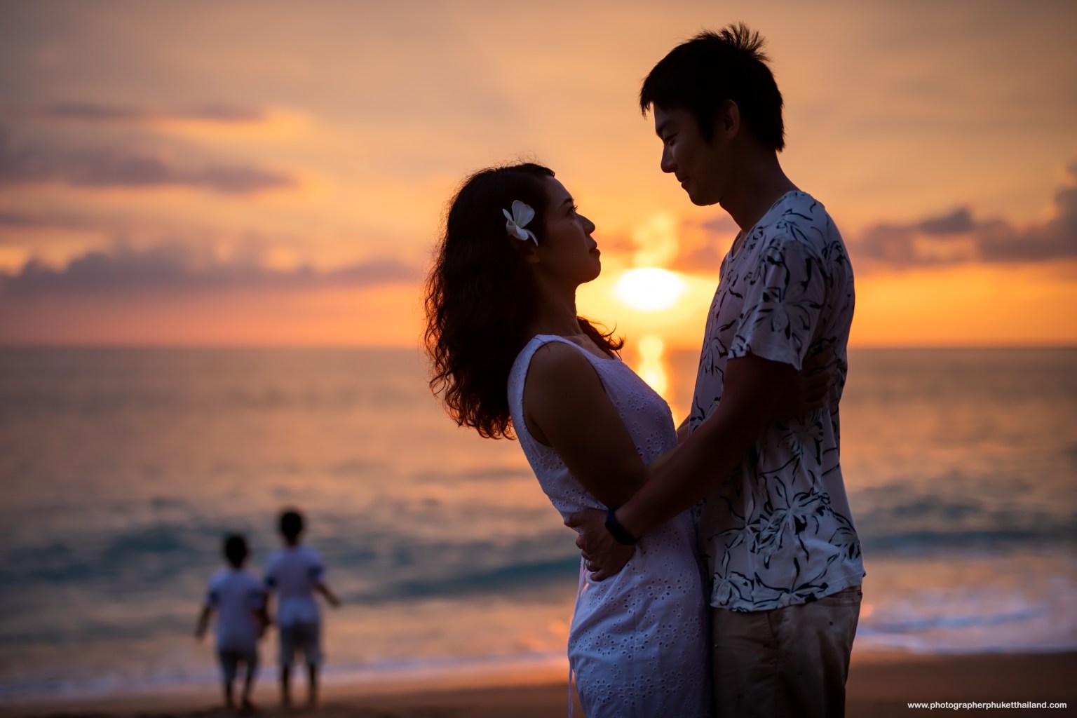 A couple embraces on the beach as sunset at mai khao beach phuket, with a beautiful orange and purple sky in the background. Two children stroll along the shore in the distance.