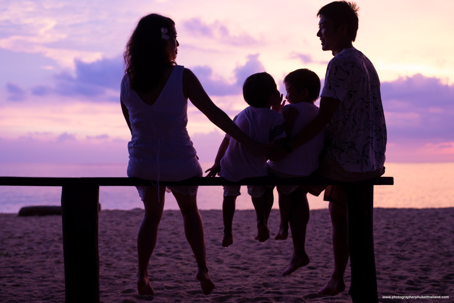 Silhouette of a family sitting on a wooden bench at the beach during sunset, with a vibrant purple sky.