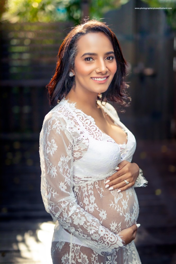 A smiling pregnant woman wearing a lace dress, standing outdoors with a blurred background, showcasing her baby bump.