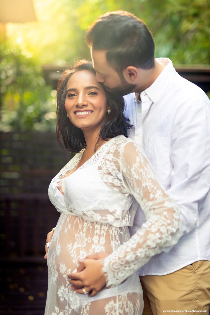 A couple poses together in a lush outdoor setting, with the woman cradling her baby bump while smiling at the camera. The man stands behind her, leaning in for a close embrace. Both are dressed in light-colored attire, with the woman wearing a lace gown.