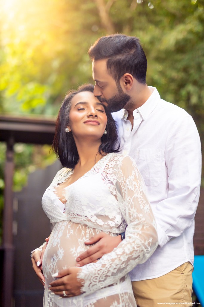 A couple embracing during a maternity photo shoot, with the woman showing her baby bump. The setting is outdoors with soft natural lighting and greenery in the background.