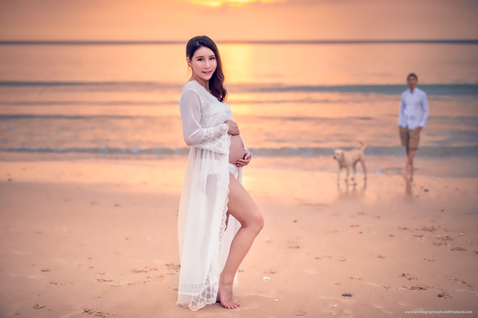 A pregnant woman in a flowing white dress poses on the beach at sunset, holding her belly, with a man and a dog walking in the background.