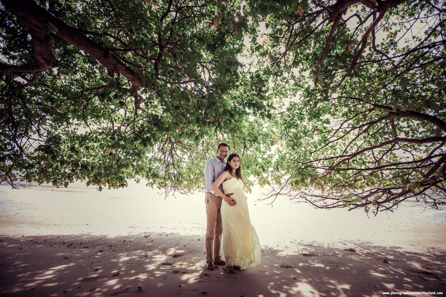 A couple posing under a large tree on the beach, with the woman in a flowing light dress and the man in a button-up shirt, showcasing a serene and romantic atmosphere.