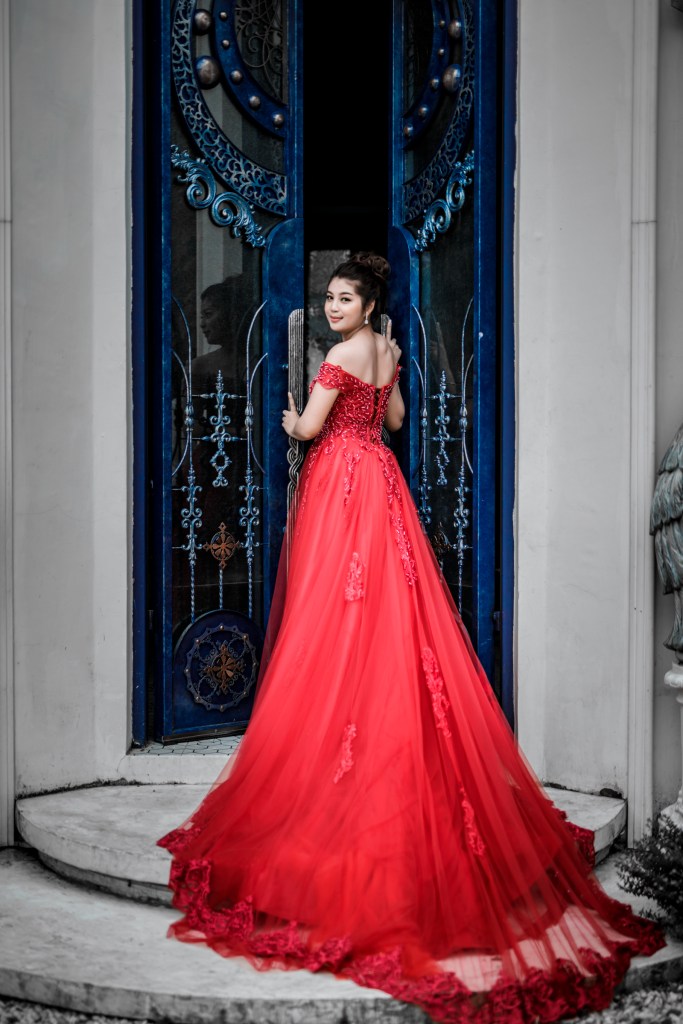Bride in a red gown posing at a blue door, showcasing her elegant hairstyle and the intricate details of her dress.