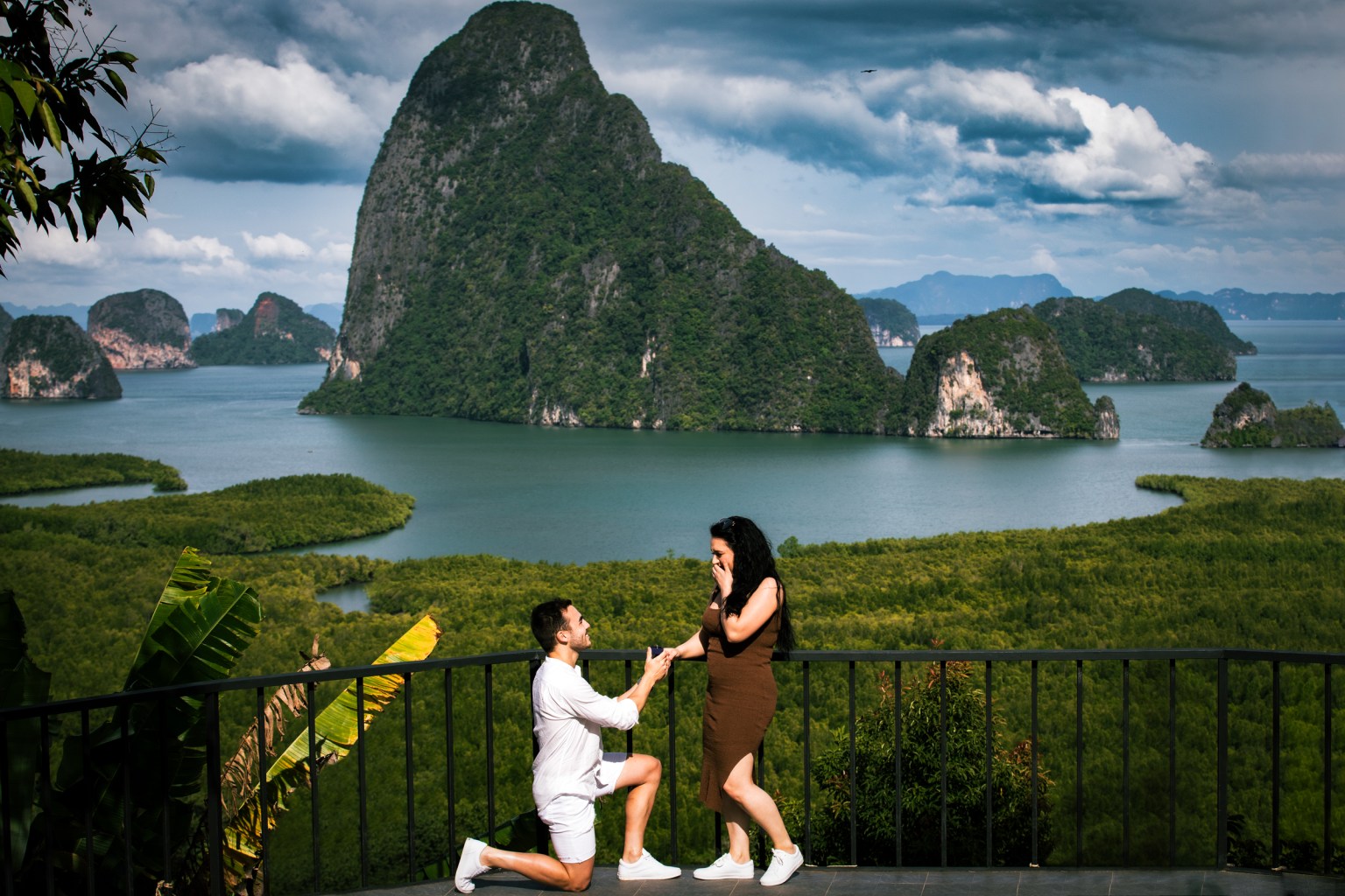 A couple on a terrace overlooking a scenic landscape at samet nangshe phang-nga bay thailand , with one partner kneeling and proposing, while the other is surprised and emotional.