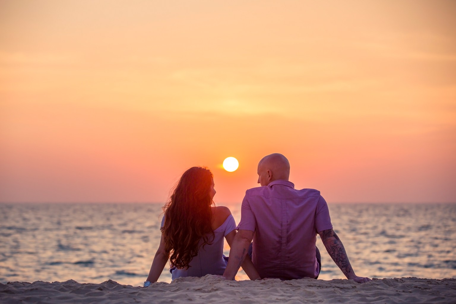 A couple sitting on the beach at sunset, looking at the ocean with a vibrant sky in the background.