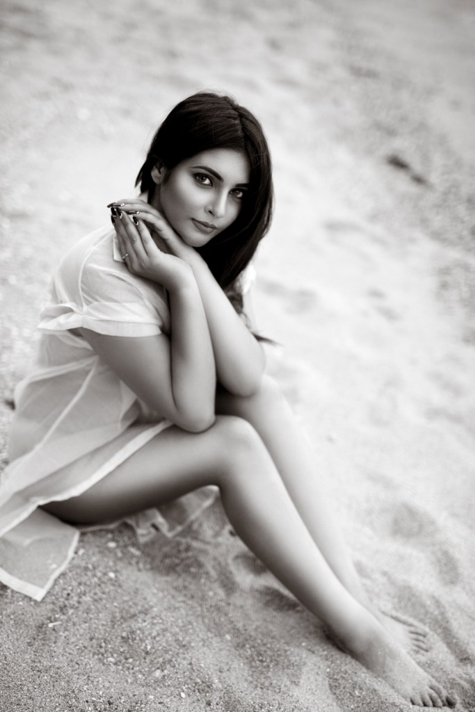 Black and white portrait of a woman sitting on the beach, with her knees drawn up and hands clasped under her chin.