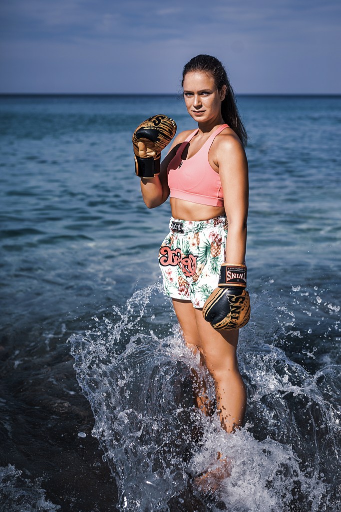 A woman stands in shallow water at the beach, wearing a pink sports top and floral boxing shorts. She holds boxing gloves and looks confidently at the camera, with waves splashing around her.