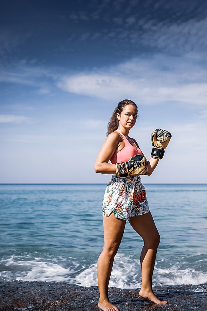 A female boxer stands confidently by the sea, wearing boxing gloves and floral shorts with a pink top, showcasing a powerful pose against a blue sky.