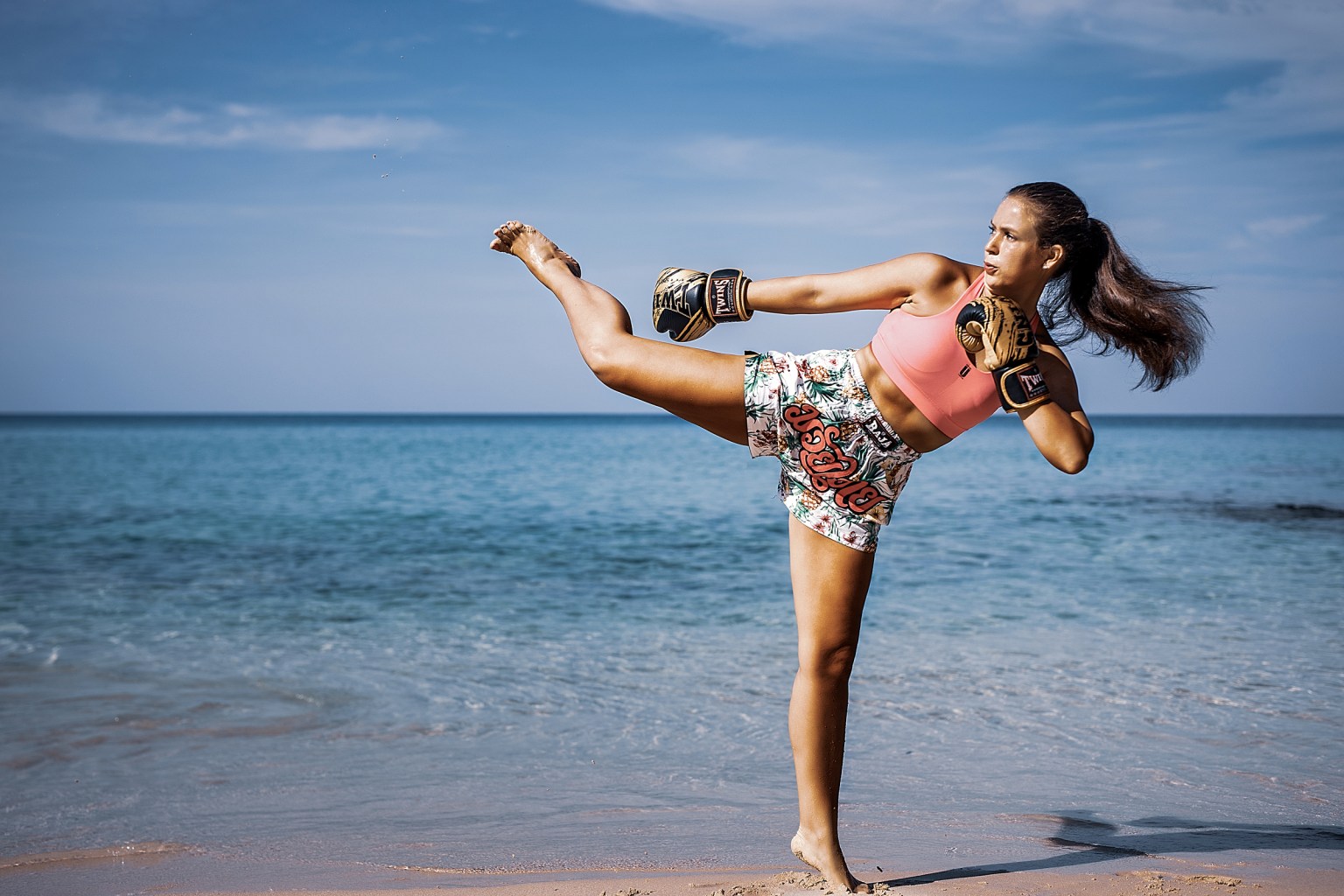 A woman performs a high kick on the beach, wearing boxing gloves and colorful shorts, against a backdrop of the ocean and clear blue sky.
