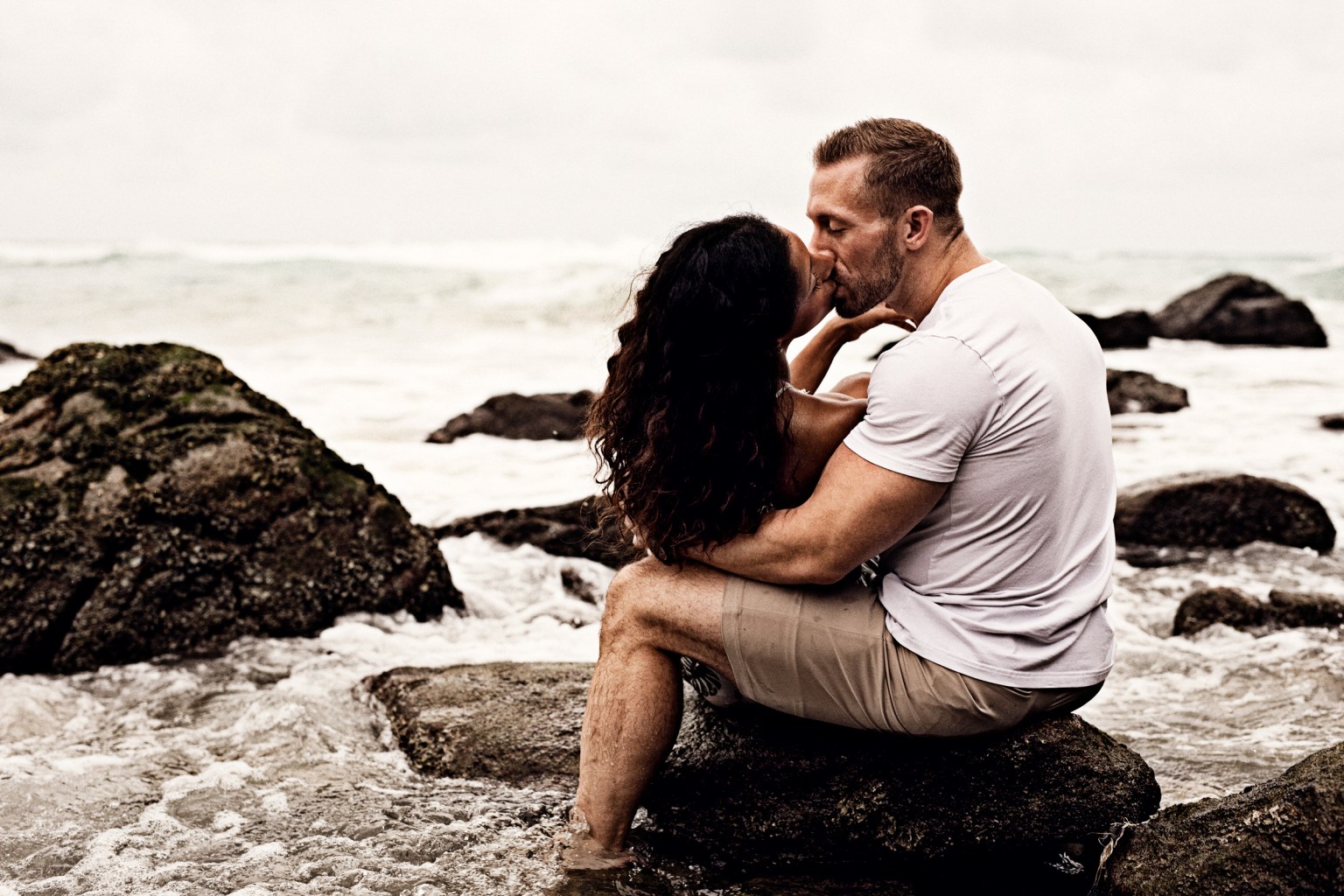 A couple sharing a romantic kiss while sitting on rocks at a beach, with waves in the background.
