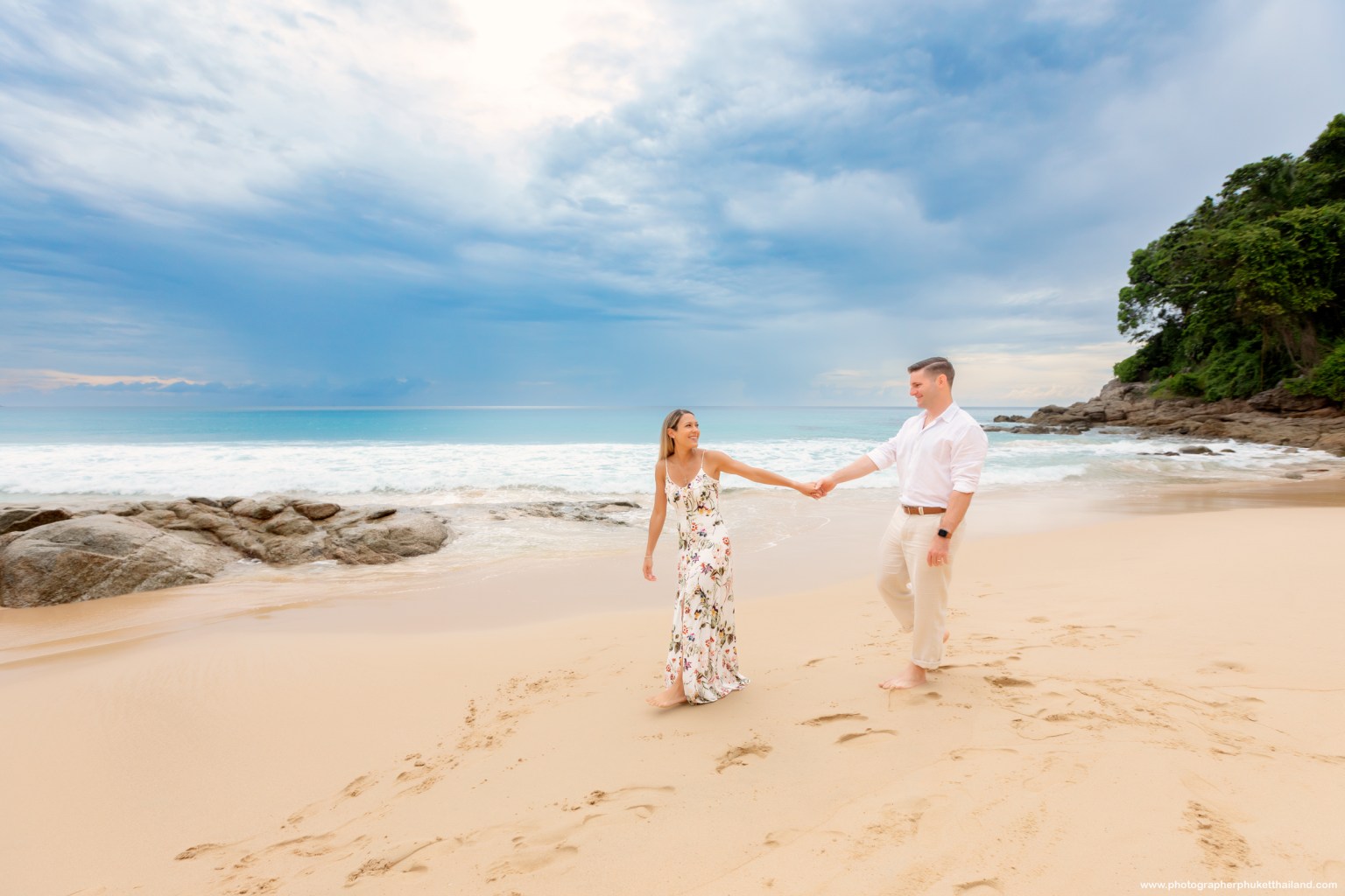 A couple walking hand in hand on a sandy beach near the ocean, with rocky formations and a cloudy sky in the background.